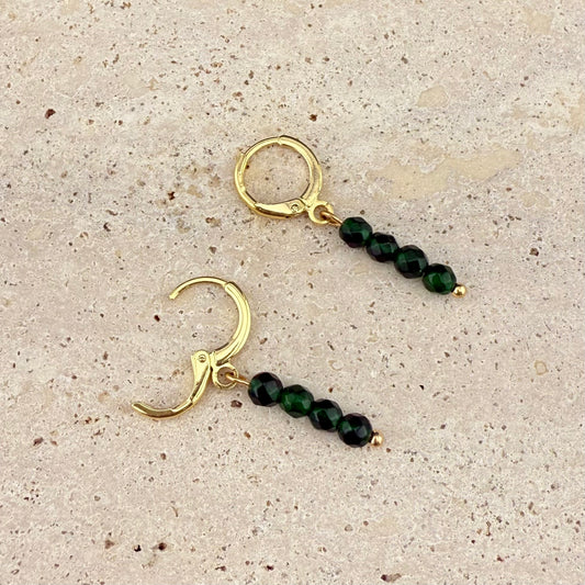 A pair of Aking Sinta gold huggie earrings with 4-bead Green Jade charms on a white textured stone; one gold clasp is open to show the easy-to-use hinge, while the other is closed, displaying the variegated green tones of the stacked beads.