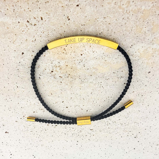 An Aking Sinta woven cord friendship bracelet in black featuring a curved gold bar. The bar is custom-engraved with the phrase "TAKE UP SPACE" in a minimalist font. The adjustable bracelet includes gold-toned accents and a branded slider, shown on a light-colored stone surface