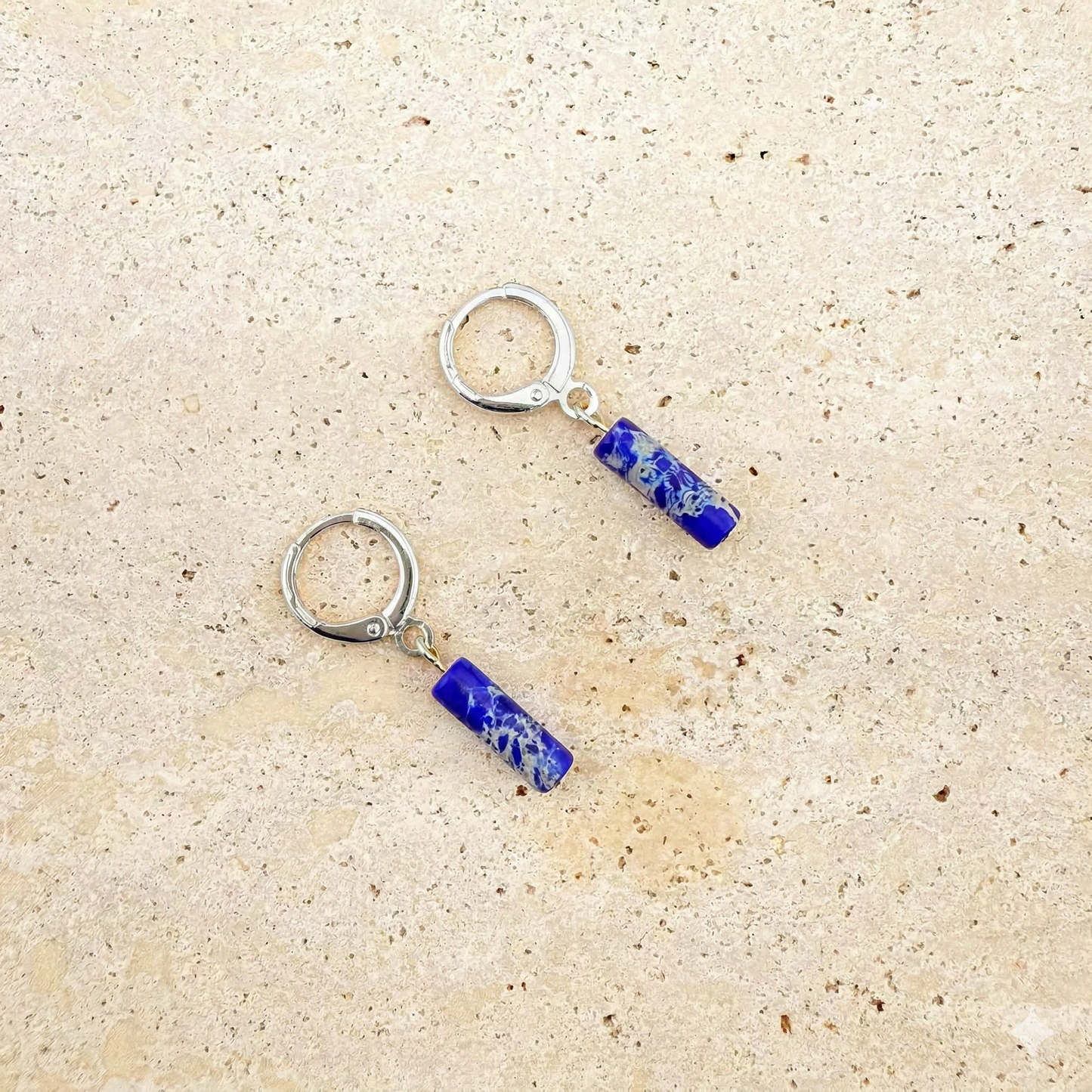 A pair of Aking Sinta silver huggie hoop earrings featuring brilliant blue imperial jasper column pendants. The marbled blue gemstone drops are displayed on a light sand-colored stone surface.