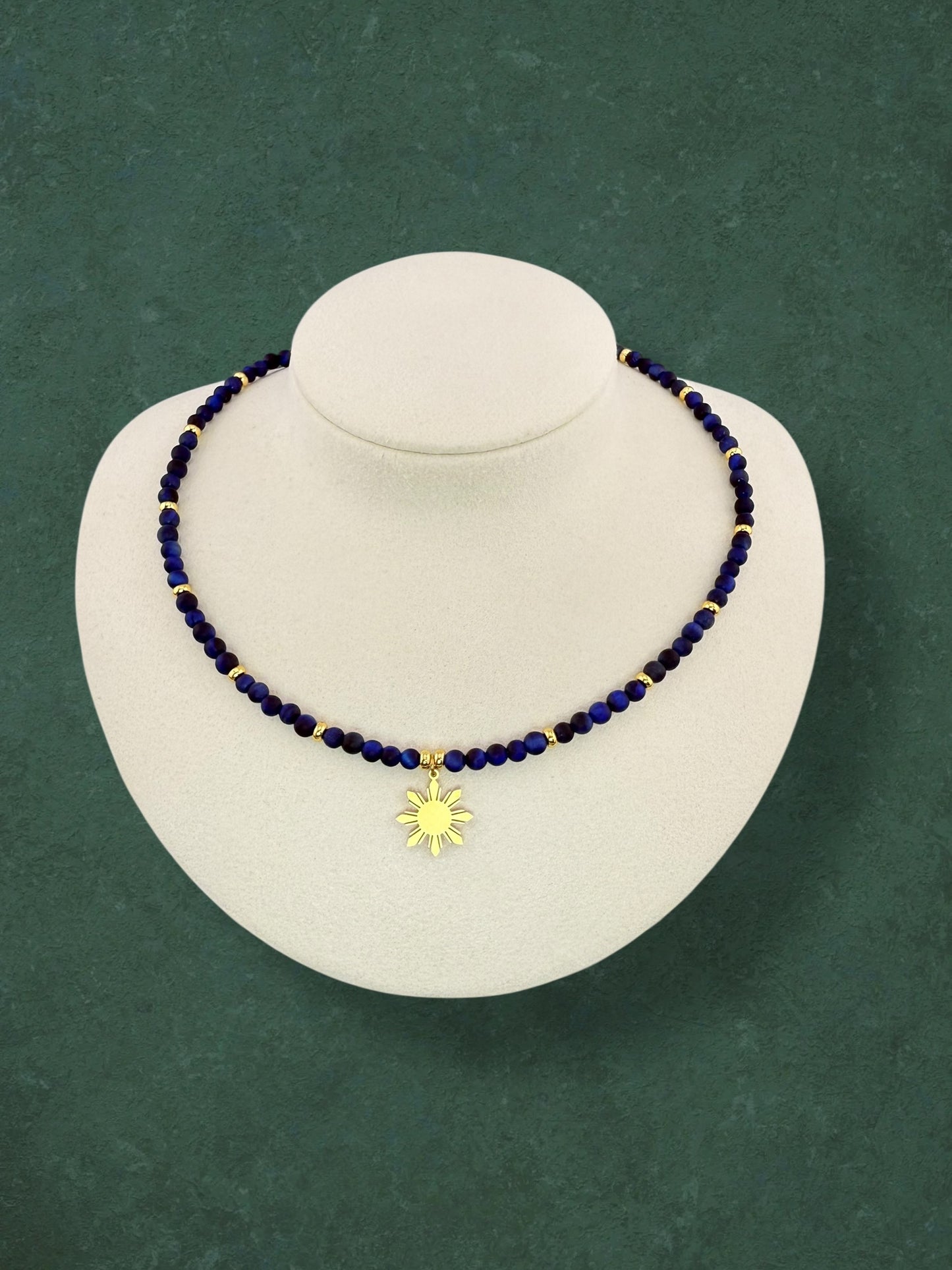 Close-up of an Aking Sinta Blue Tiger Eye beaded necklace with a central gold 8-ray Araw sun charm; the dark blue stones show a cat-eye shimmer against a solid dark green background.