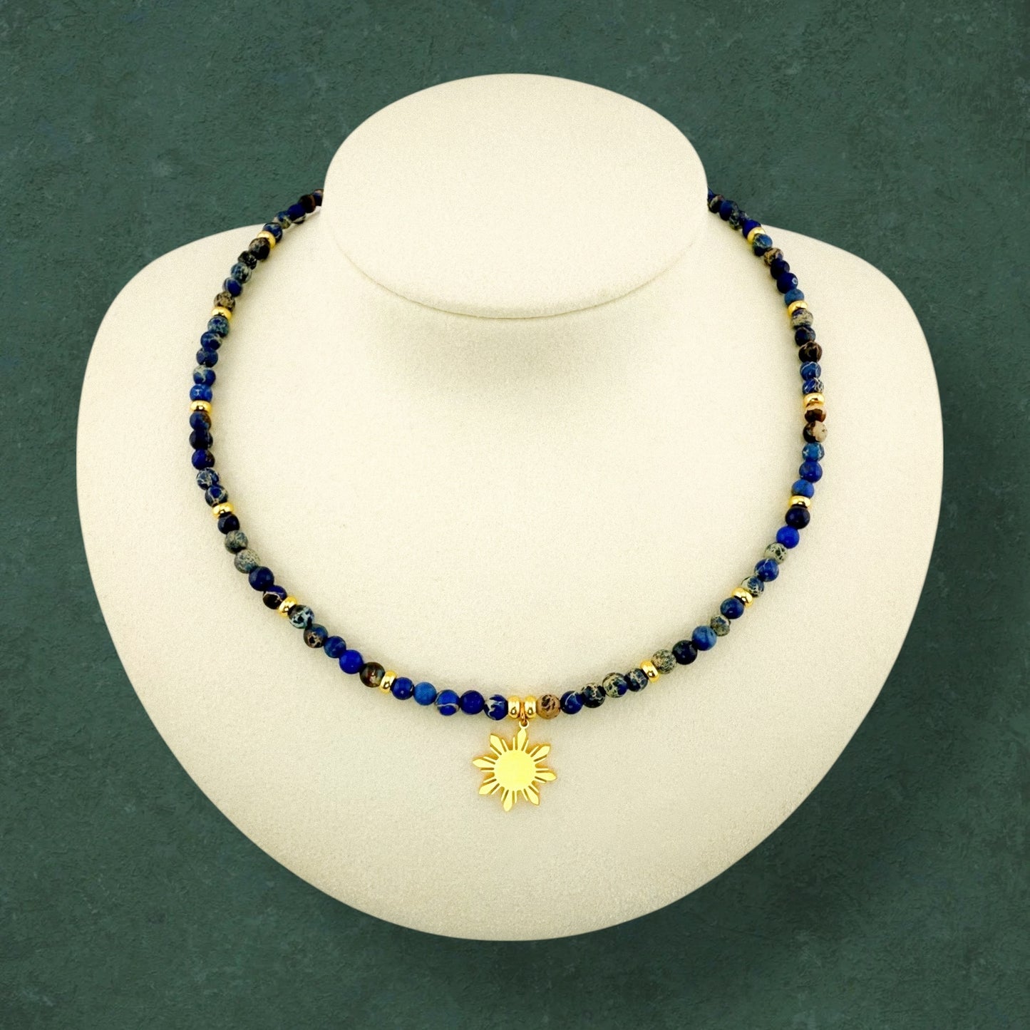 Aking Sinta Brilliant Blue Imperial Jasper necklace featuring gold bead accents and a central 8-ray Araw sun charm; the vibrant blue stones are displayed on a dark green background.