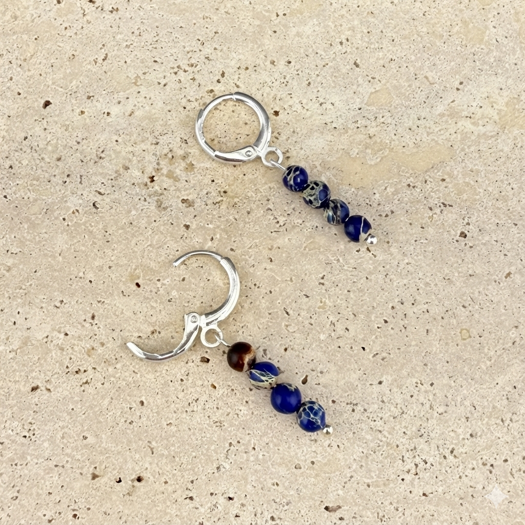 Silver variant of Aking Sinta Brilliant Blue Imperial Jasper huggies on a gray rock-like surface; one silver hinge clasp is shown open to reveal the mechanism and the vibrant blue gemstone beads.