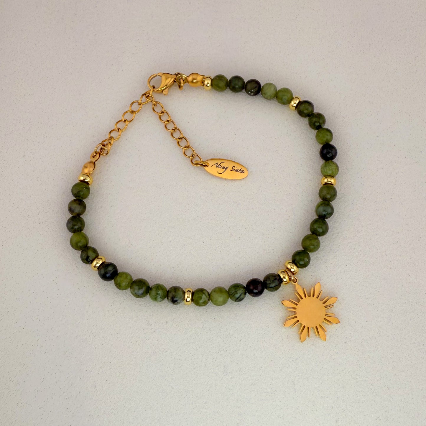 An Aking Sinta Canadian Jade beaded bracelet featuring smooth, translucent forest-green gemstones and a signature gold 8-ray Araw sun charm, displayed on a clean white background