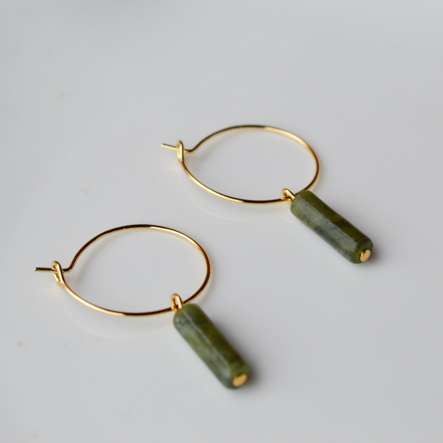 A pair of gold hoop earrings with vertical Canadian Jade column charms resting inside a shimmering Mother of Pearl shell container; the dark green translucent stones contrast beautifully with the iridescent pink and white background.
