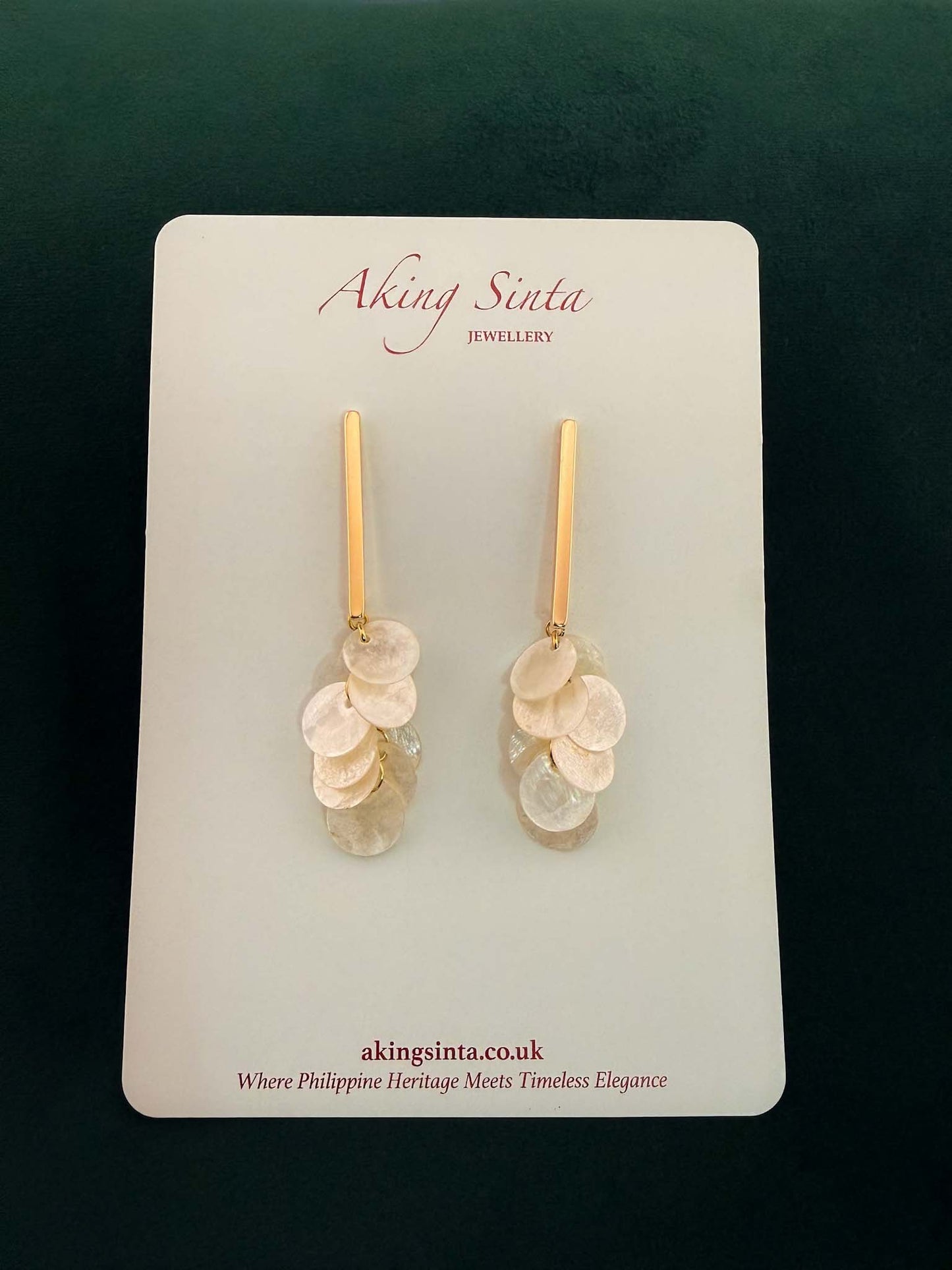 Pair of gold Aking Sinta cascading Capiz shell earrings on bars, set against a dark forest green velvet background
