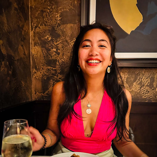 A candid, smiling portrait of a woman wearing an Aking Sinta gold sun motif and Capiz shell pendant necklace with matching drop earrings. She is dressed in a vibrant pink halter top, captured in a warm, dimly lit restaurant setting, showcasing how the jewelry styles for an evening ou
