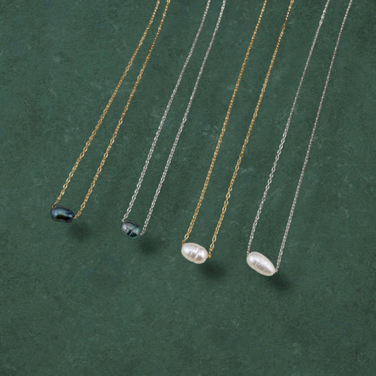 A curated flat-lay of Aking Sinta floating pearl necklaces, showcasing four variations featuring lustrous white and iridescent dark pearls strung on delicate gold and silver chains. The necklaces are elegantly arranged in parallel on a rich, deep green textured background, highlighting the minimalist and versatile design of the collection