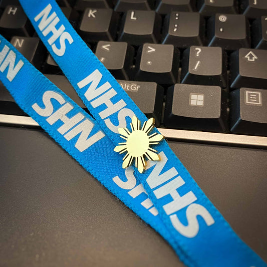 A gold Aking Sinta Araw lapel pin with an 8-ray sunburst design attached to a blue ID lanyard; a computer keyboard is visible in the blurred background, showcasing the pin as a stylish office accessory