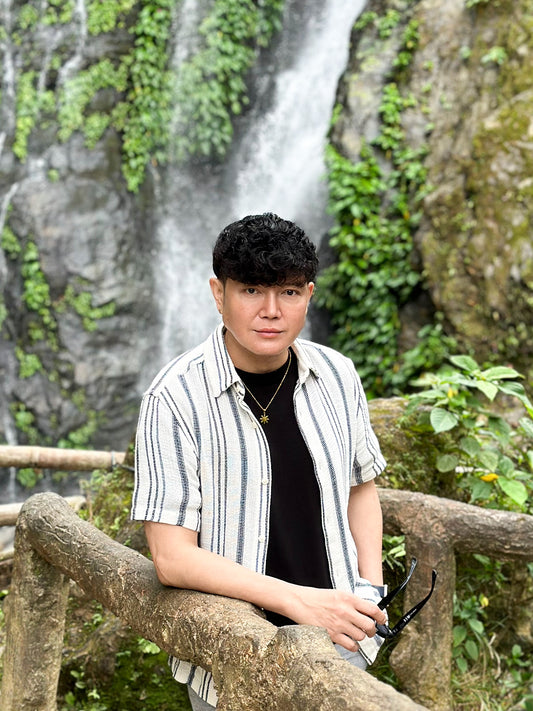 A male lifestyle shot featuring the Aking Sinta gold Araw necklace. The model wears the minimalist Philippine sun pendant over a black t-shirt and striped button-down shirt, standing against a lush, tropical waterfall background. The image highlights the necklace as a versatile, unisex piece of Filipino-inspired heritage jewelry.