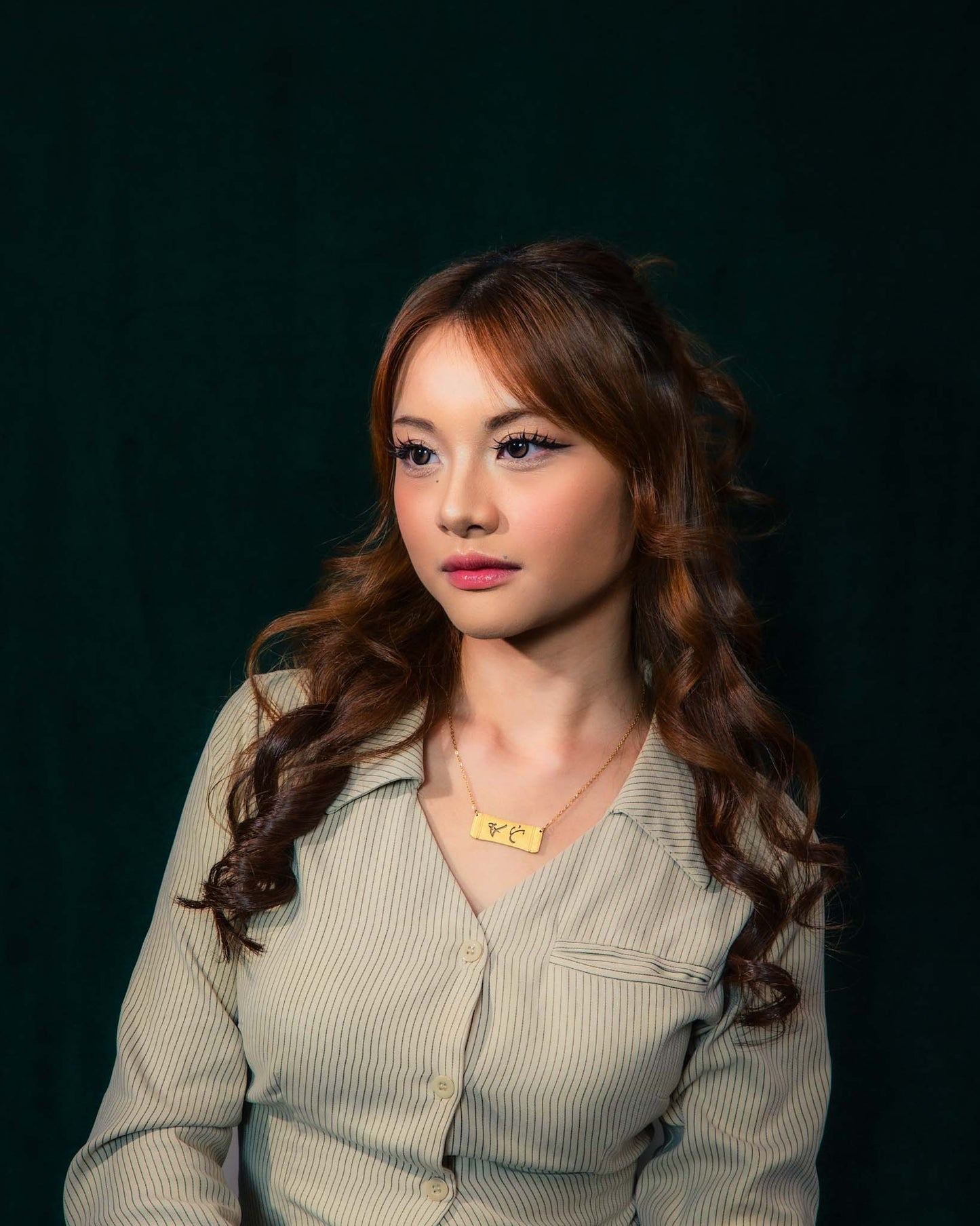 A studio lifestyle portrait of a woman with long, curly brown hair wearing the Aking Sinta gold Ate bamboo bar necklace. The horizontal pendant featuring the Baybayin script for elder sister is styled over a pinstriped beige button-down shirt against a dark green studio backdrop