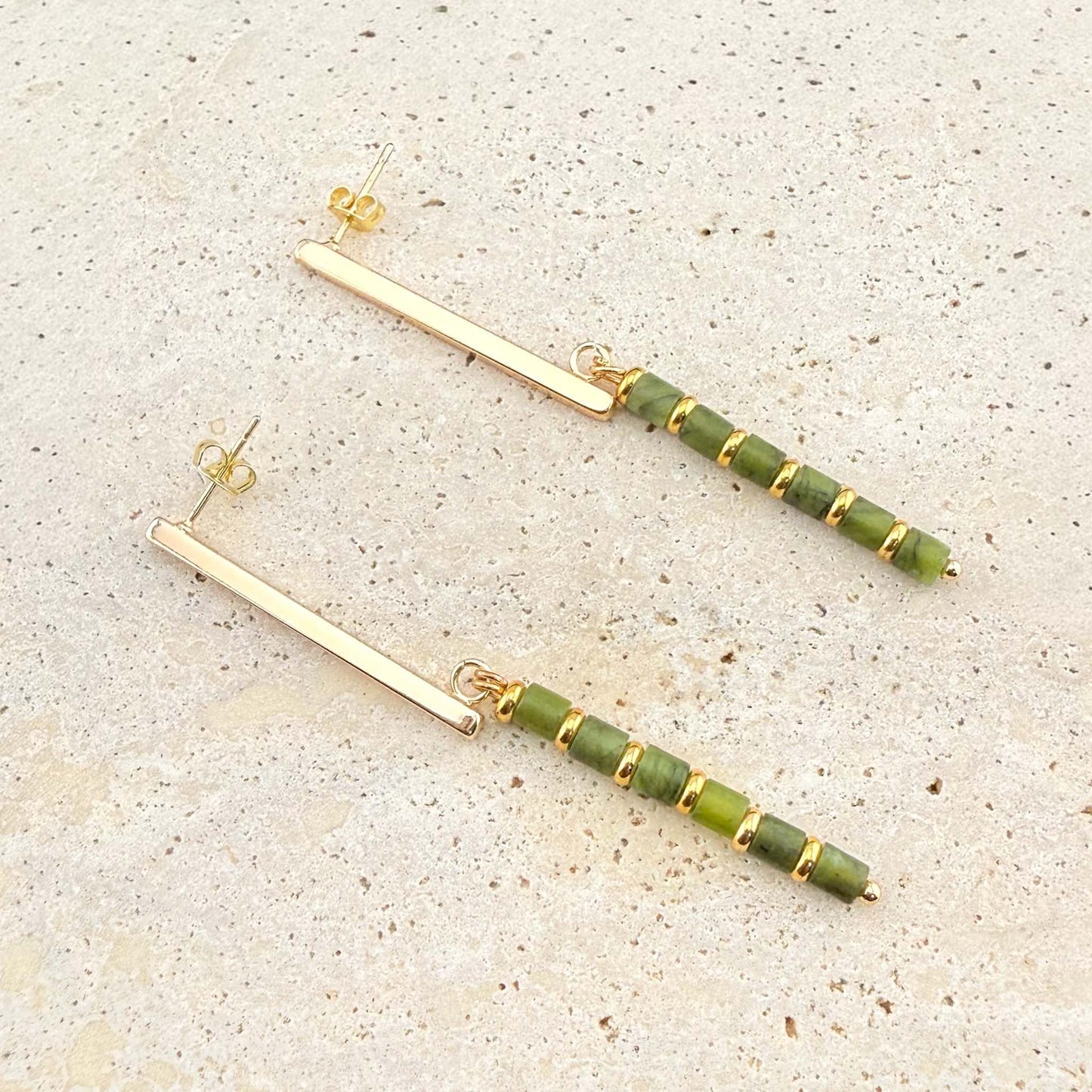 A pair of elegant Aking Sinta gold bar drop stud earrings. Each earring features a sleek, polished gold bar stud from which a long drop hangs, composed of alternating small, round Canadian jade beads and gold spacers. The earrings are displayed on a light-colored natural stone surface