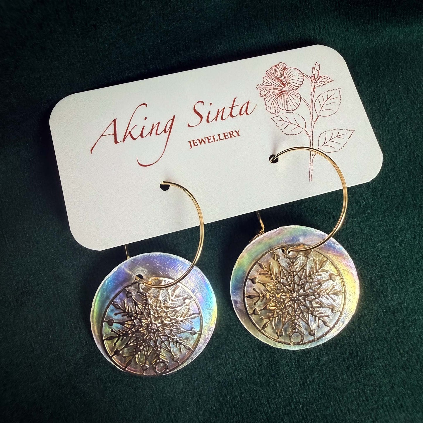 A pair of Aking Sinta gold hoop earrings featuring large, iridescent Capiz shell discs layered with intricate gold snowflake charms. The earrings are displayed on a white branded jewelry card against a rich, dark green velvet background, highlighting the shimmering rainbow luster of the shells and the fine metallic detail of the snowflakes