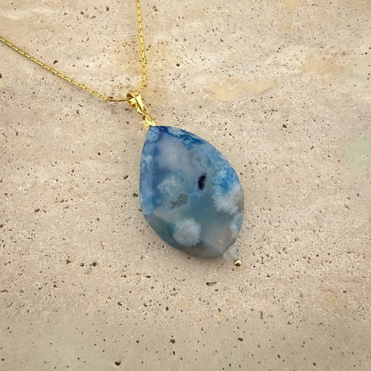 A detailed close-up of an Aking Sinta gold chain necklace featuring a faceted teardrop-shaped blue agate pendant. The gemstone displays intricate, cloud-like white inclusions against a soft blue backdrop, elegantly presented on a light, textured natural stone surface