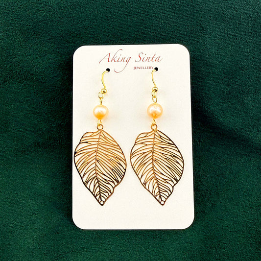 A pair of Aking Sinta gold filigree leaf and peach pearl drop earrings on a white branded display card. The delicate, nature-inspired earrings are set against a rich dark green velvet background.