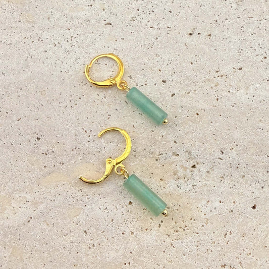 A pair of Aking Sinta gold huggie hoop earrings with green aventurine column drops. One earring is open to show the clasp mechanism, presented on a natural light-colored stone background