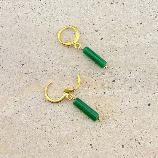 A pair of Aking Sinta gold huggie earrings with deep green jade column pendants. One earring is unlatched to show the hinge clasp design on a light-colored natural stone background