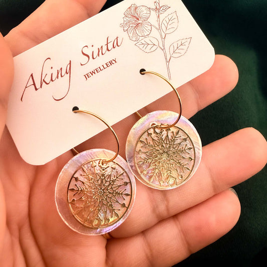 A close-up shot of a hand holding a pair of Aking Sinta gold hoop earrings on a branded jewelry card. The earrings feature luminous, iridescent round Capiz shell discs layered with intricate gold snowflake charms, showcasing the delicate craftsmanship and shimmering textures of the Filipino-inspired design