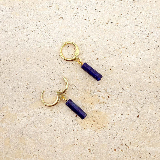 A pair of gold Aking Sinta huggie hoop earrings featuring deep blue lapis lazuli column charms. One hoop is shown with the clasp open, showcasing the hinge design. The natural gemstone pendants feature a rich blue color and hang from polished gold-toned hoops on a light natural stone surface
