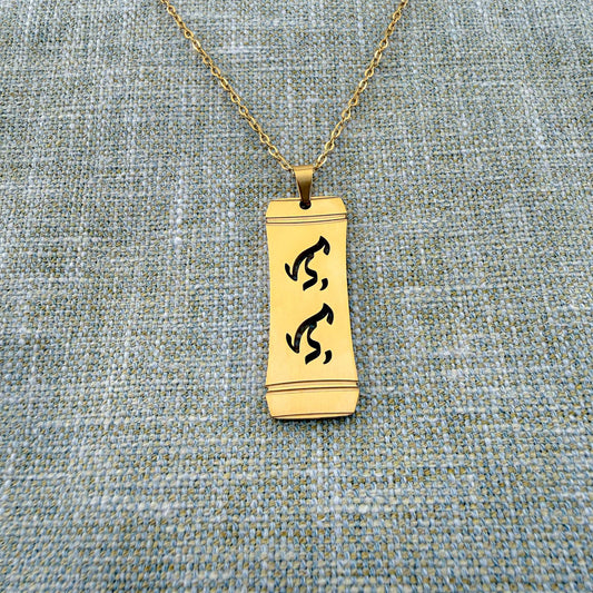 An Aking Sinta gold vertical pendant necklace centered on a textured blue-gray woven linen backdrop. The matte gold rectangular pendant features a stylized bamboo frame outline with the word "Lolo" (Grandfather) intricately cut out in traditional script-like Baybayin characters, hanging from a fine gold link chain