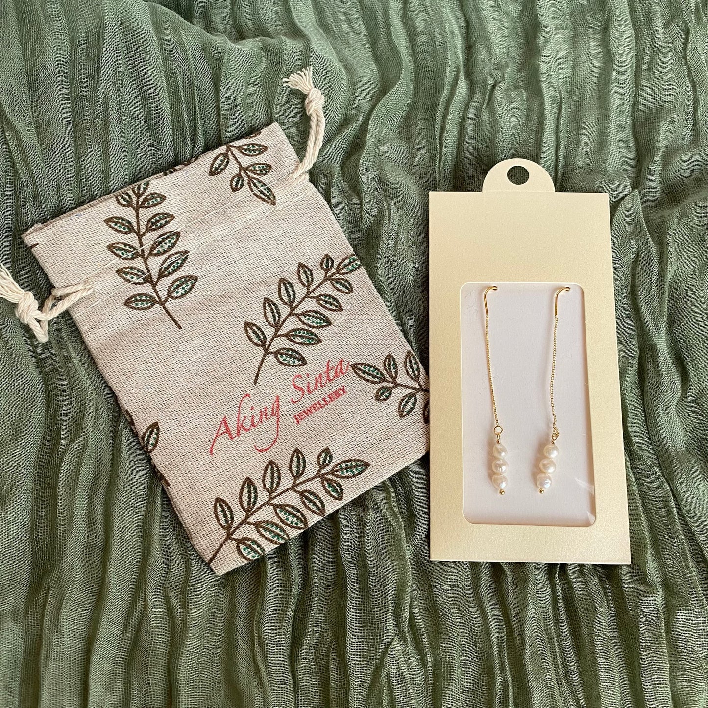 A pair of dainty Aking Sinta gold threader earrings accented with small pearls, presented in branded cream-colored packaging. Beside the earrings lies a matching Aking Sinta jewelry drawstring pouch, featuring a delicate leaf pattern. The items are laid out on a soft, textured olive green fabric, showcasing the brand's elegant, gift-ready presentation