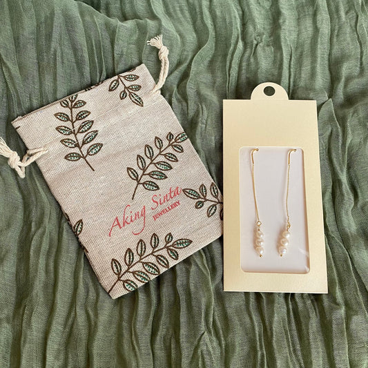 A pair of dainty Aking Sinta gold threader earrings accented with small pearls, presented in branded cream-colored packaging. Beside the earrings lies a matching Aking Sinta jewelry drawstring pouch, featuring a delicate leaf pattern. The items are laid out on a soft, textured olive green fabric, showcasing the brand's elegant, gift-ready presentation