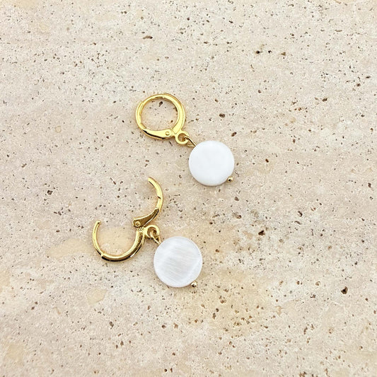 A pair of Aking Sinta gold huggie hoop earrings with circular mother of pearl disc charms. One earring is open to show the clasp, highlighting the iridescent white shell drops on a light stone background.
