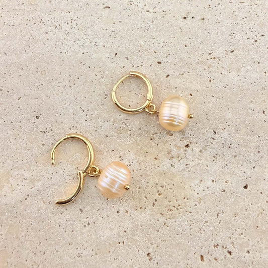 A pair of Aking Sinta gold huggie hoop earrings with peach freshwater pearl drops. One earring is unlatched to show the hinge closure, highlighting the lustrous round pearls on a light-colored natural stone background.