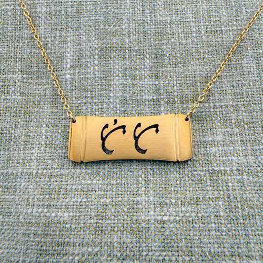 An Aking Sinta gold horizontal bar necklace centered on a textured blue and gold woven linen background. The matte gold pendant features a stylized bamboo section silhouette with the word "Tita" (Aunt) cleanly cut out in elegant, traditional Baybayin script characters, suspended from a fine gold link chain.