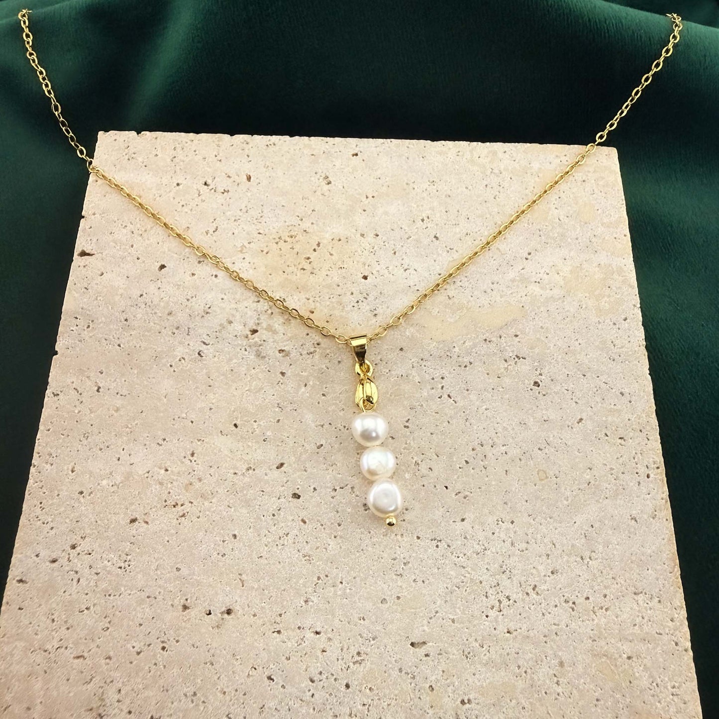 A gold Aking Sinta necklace featuring a delicate pendant of three vertically stacked, organic white baroque pearls. The pendant is suspended from a polished gold bail and fine cable chain, artfully displayed on a light, textured stone block against a rich dark green velvet background