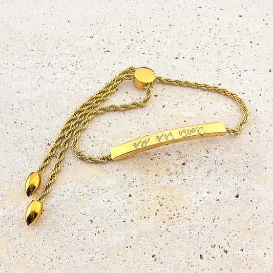 A gold Aking Sinta twisted rope chain bracelet with a bespoke engraving. The curved gold bar is etched with ancient Filipino Baybayin script and features an adjustable bolo-style slider clasp with teardrop-shaped end charms, displayed on a light natural stone surface