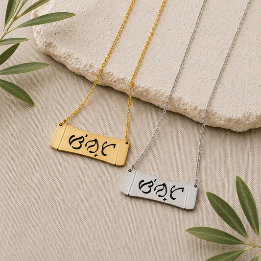 A product lifestyle photograph featuring two Aking Sinta horizontal bamboo bar necklaces in matte gold and silver finishes. The parallel curved bar pendants showcase the brand's namesake word "Sinta" (Beloved) beautifully cut out in traditional script-like Baybayin characters, arranged on a cream-colored textured woven surface next to a light stone slab and fresh olive leaves.