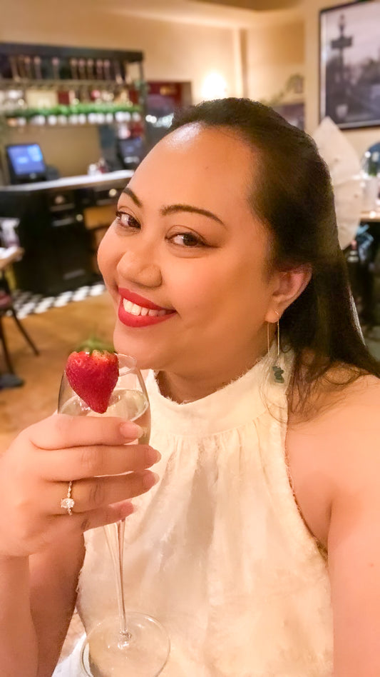 Lifestyle photo of a woman in a white dress wearing Aking Sinta gold drop earrings with clustered green aventurine nuggets; she is holding a champagne glass against a dark green background, highlighting the jewelry's elegant and celebratory appeal.