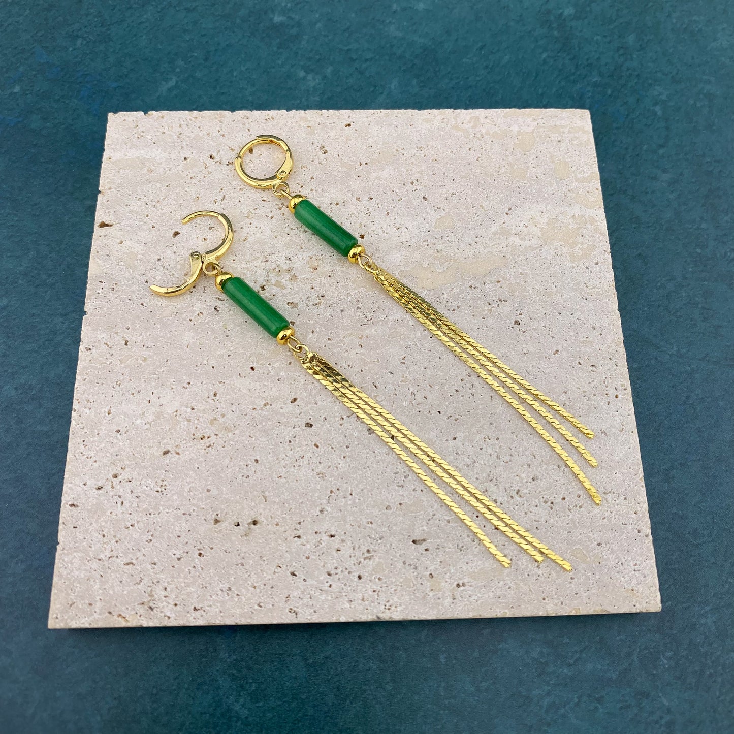 Macro view of a Green Jade column tassel earring on gray marble with the gold huggie clasp open, showing the internal hinge and the detailed attachment of the jade stone and gold chain fringe.