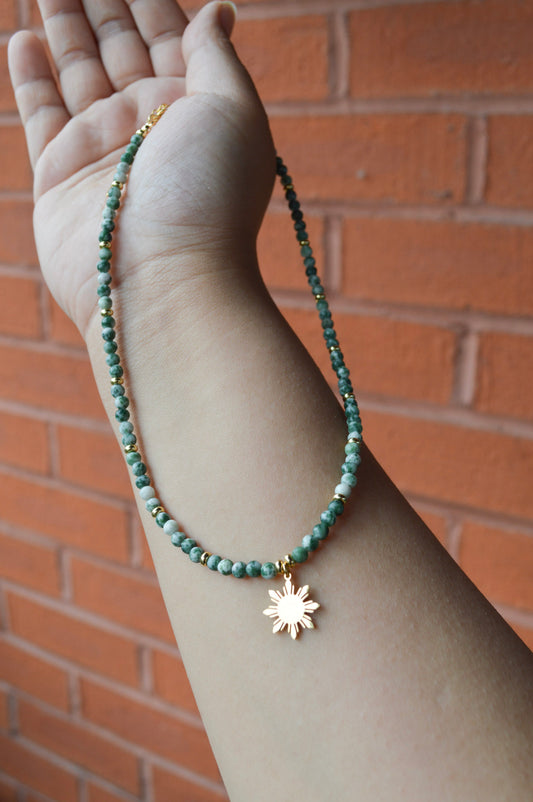 A Green Spot Jasper stone necklace with a gold 8-ray Araw charm displayed on a model's palm; the hand is held in an open, receiving gesture against a blurred background, highlighting the earthy tones of the Aking Sinta jewelry.