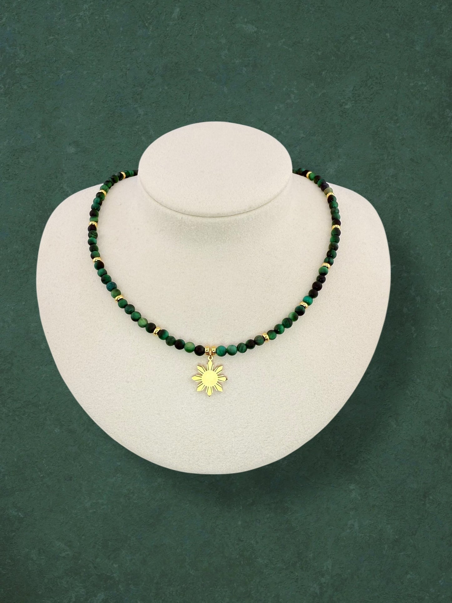 Aking Sinta Green Tiger Eye beaded necklace displayed on a white bust; featuring deep emerald-green gemstones with a silky, light-catching shimmer, accented with polished gold beads integrated into the strand and a central gold 8-ray Araw sun charm