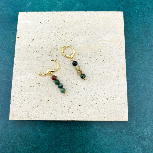 A pair of gold Aking Sinta huggie earrings with round Indian Agate gemstone charms, featuring natural swirls of forest green and earth brown, resting on a large, polished gray marble slab.