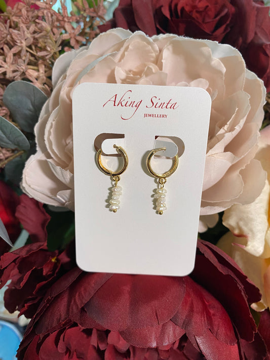 Macro close-up of a single Aking Sinta Stacked Keshi Pearl Huggie earring on a branded card; showing the detailed iridescent texture of the organic pearls against a blurred rose background.