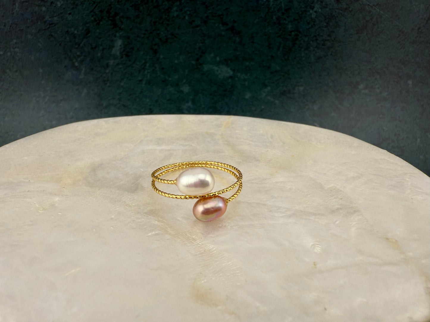 Birds-eye view of an Aking Sinta gold adjustable ring with lilac and white pearls, placed off-center on a circular white marble slab against a dark green background.