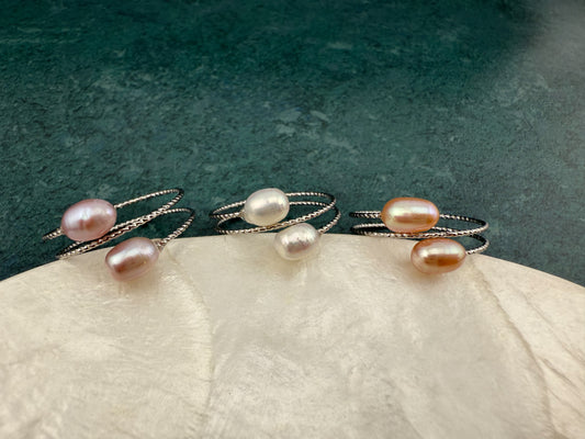 Macro close-up of three Aking Sinta double pearl rings—Peach, White, and Lilac—resting on the edge of a white marble slab against a dark green background.