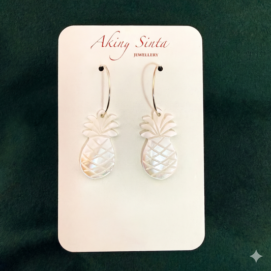 A pair of Aking Sinta pineapple hoop earrings on a white branded display card. Each earring features a carved white mother of pearl pineapple charm suspended from a silver hoop, set against a dark green textured background.
