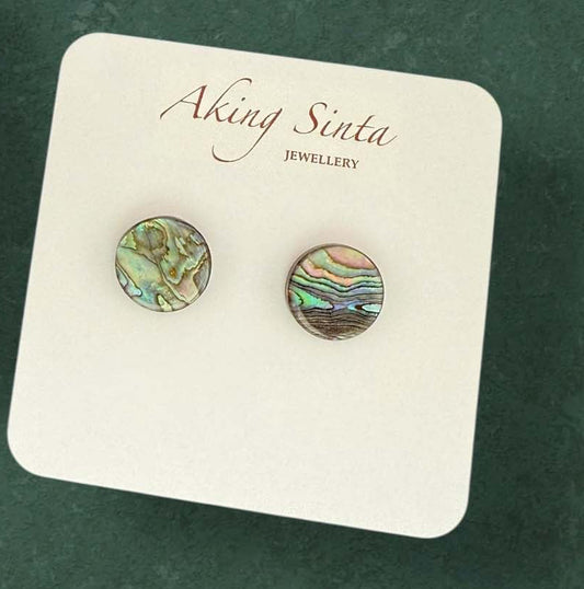A pair of Aking Sinta small round abalone shell stud earrings. The natural iridescent shell displays swirls of ocean green, blue, and pink, set in a minimalist frame on a white jewelry card