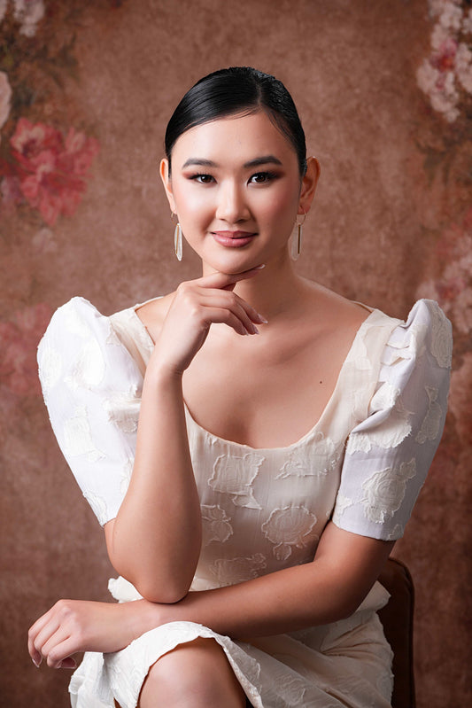 A close-up side view of a model wearing a white top and Bar Accented Oval Capiz Hoops; the model is resting her right hand on her chin, showcasing the elongated oval shell disk and its polished gold center bar.