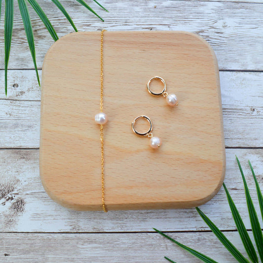 A matching Aking Sinta jewelry set featuring a peach pearl minimalist gold bracelet and gold huggie hoop earrings with pearl drops. Displayed on a wooden block with tropical palm leaf accents