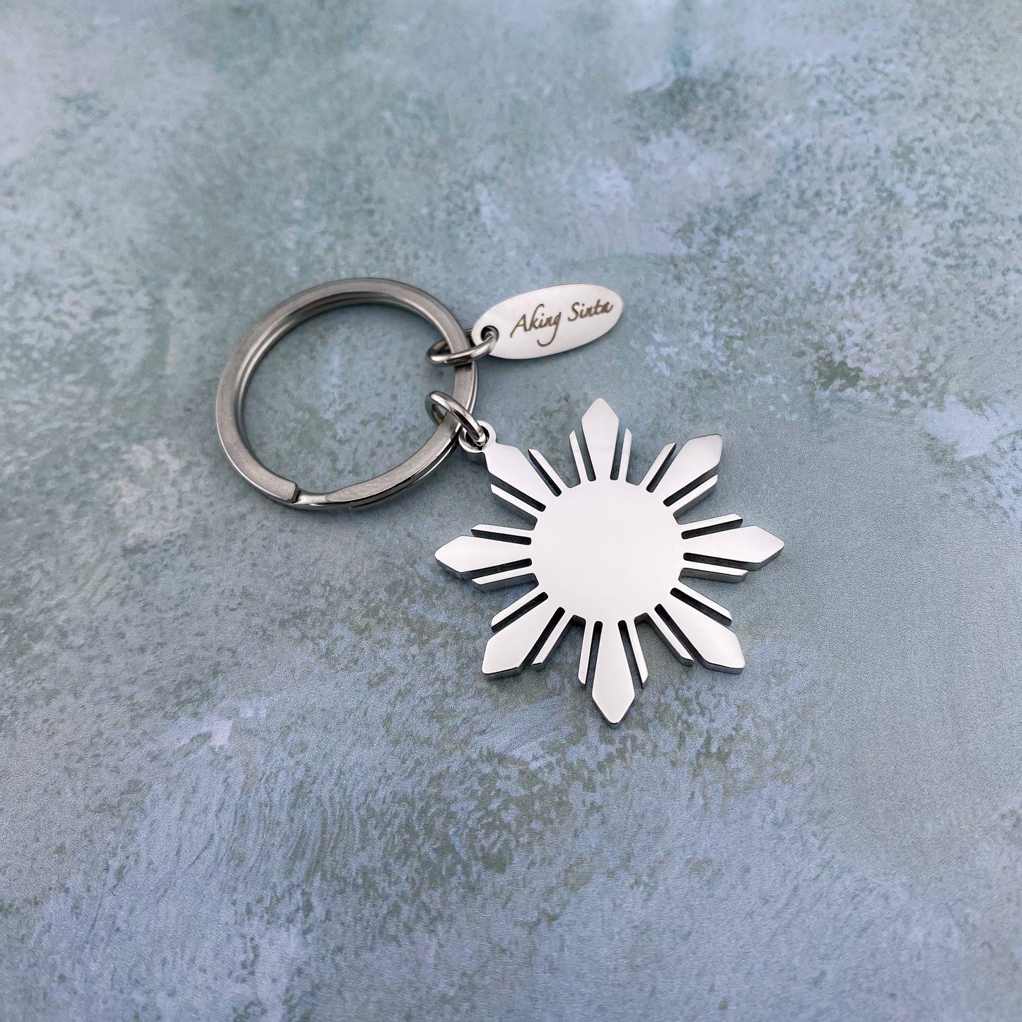 The silver Aking Sinta Araw key ring with a large 8-ray sunburst charm, elegantly placed on a gray marble background to showcase the brilliant metallic shine and durable craftsmanship