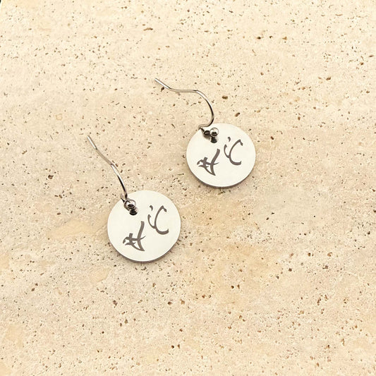 A pair of silver Aking Sinta hook earrings featuring circular coin charms with bespoke engravings. The silver discs are etched with ancient Filipino Baybayin script and hang from delicate fishhook wires, displayed on a light, textured natural stone surface