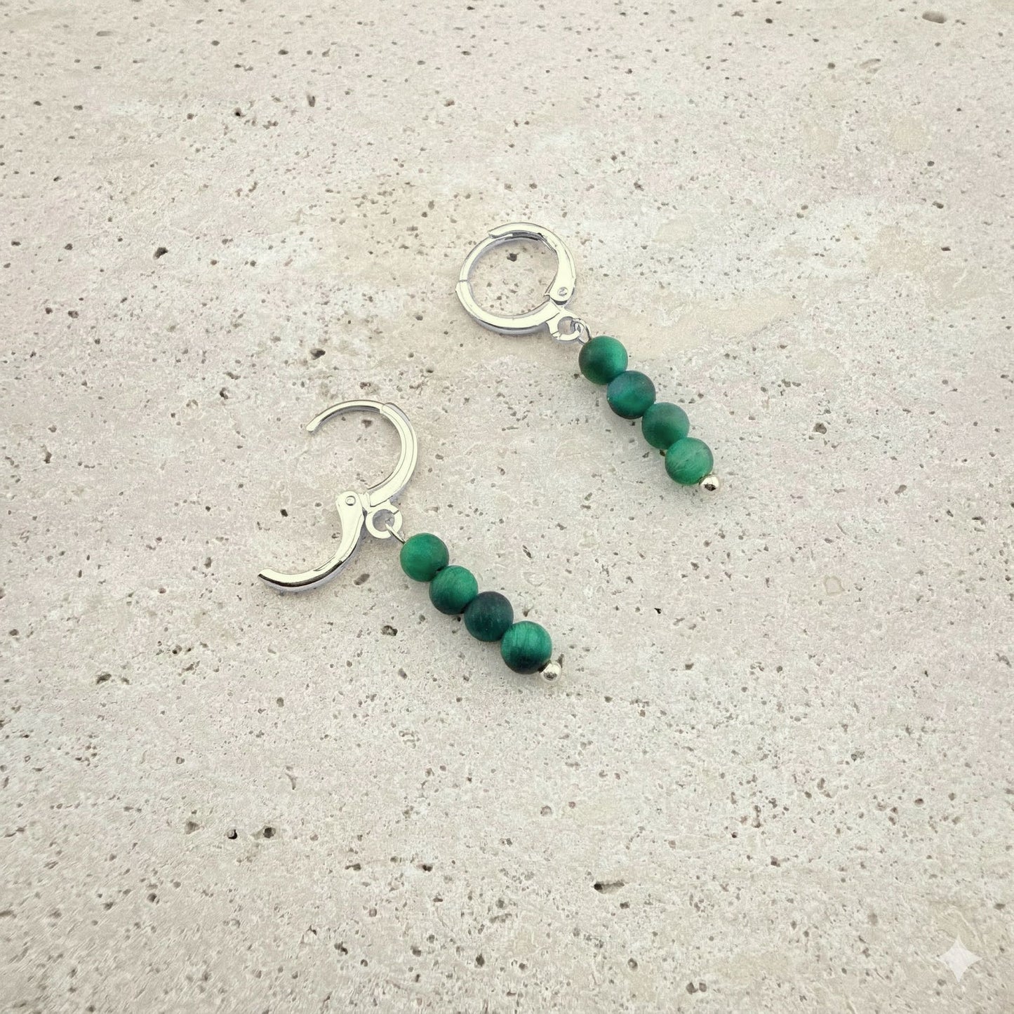 A pair of silver Aking Sinta huggie hoop earrings, each featuring a dangling drop of four small, rich green tiger eye gemstone beads. The earrings are elegantly displayed against a light, textured natural stone surface.