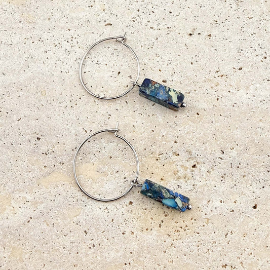 A pair of Aking Sinta silver wire hoop earrings featuring blue imperial jasper cuboid gemstone pendants. The unique, marbled blue and earth-toned stone drops are displayed on a light sand-colored stone surface