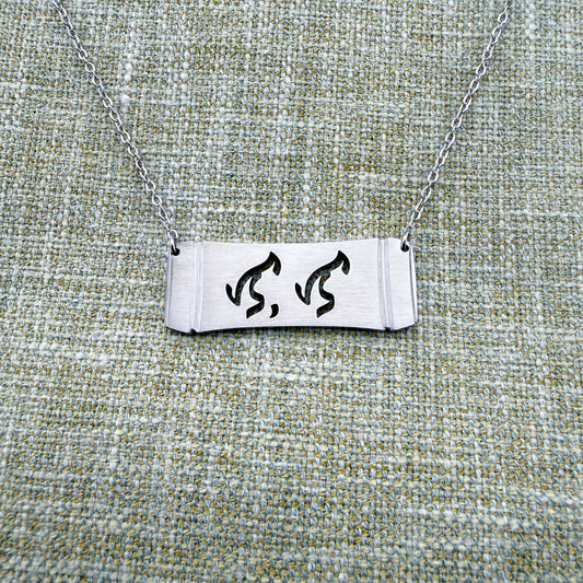 An Aking Sinta silver horizontal bar necklace centered on a textured green-gray linen background. The matte silver pendant features a curved bamboo frame silhouette with the word "Lola" (Grandmother) elegantly cut out in traditional script-like Baybayin characters, suspended by a fine silver link chain.