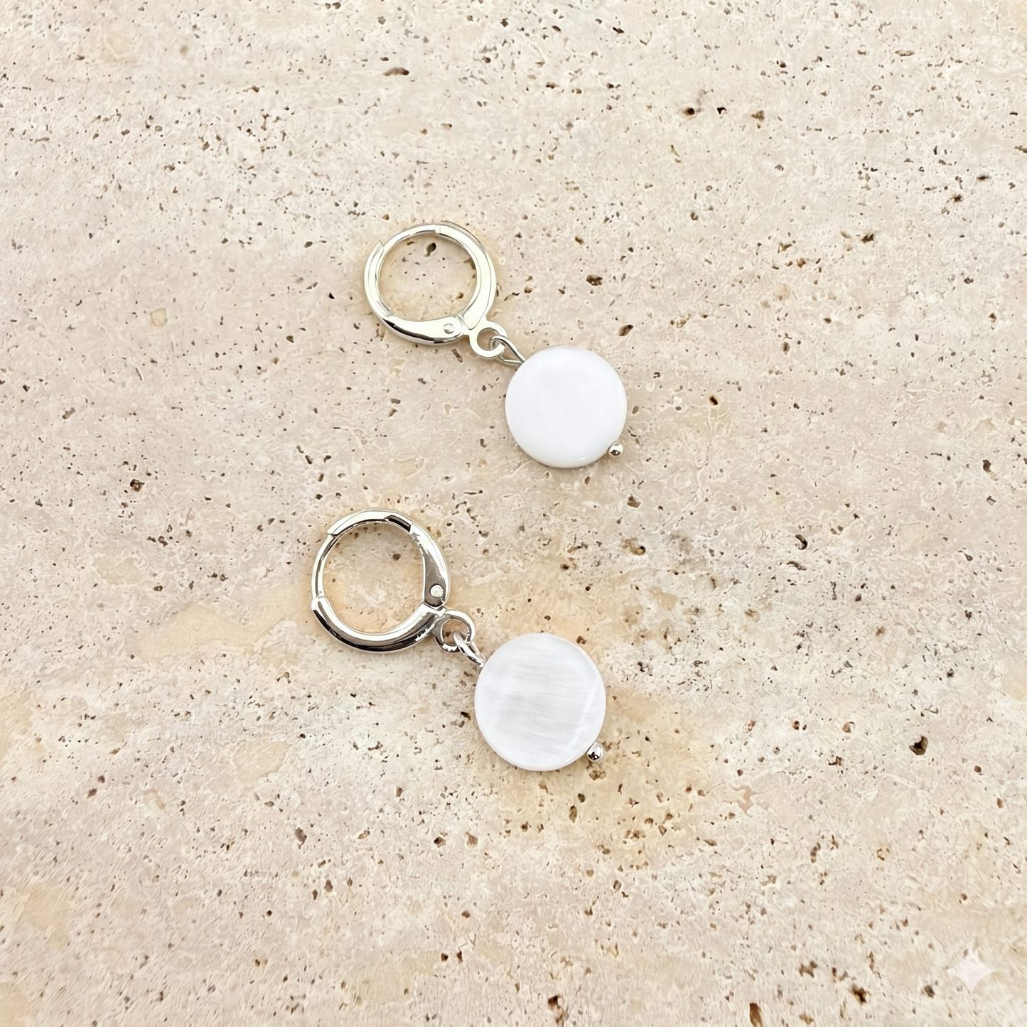 A pair of Aking Sinta silver huggie hoop earrings featuring circular mother of pearl disc charms. The iridescent white shell drops are displayed on a light, textured stone background.