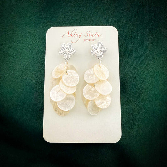 A pair of silver Aking Sinta statement earrings featuring intricate Sampaguita flower studs. Clusters of iridescent white Capiz shell discs cascade from the silver floral tops. The jewelry is displayed on a branded white card against a rich dark green velvet background